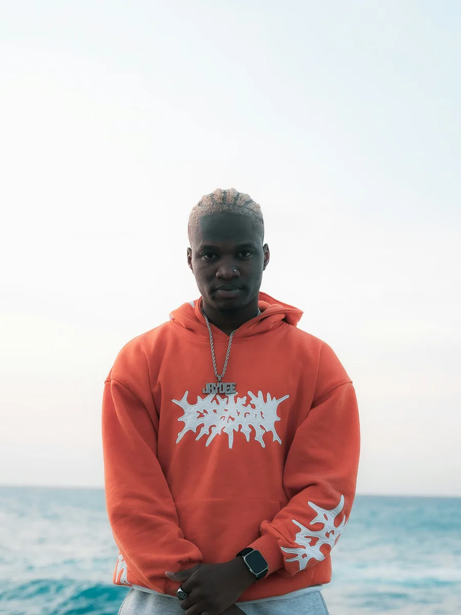 Man in an orange graphic hoodie and grey joggers by the sea